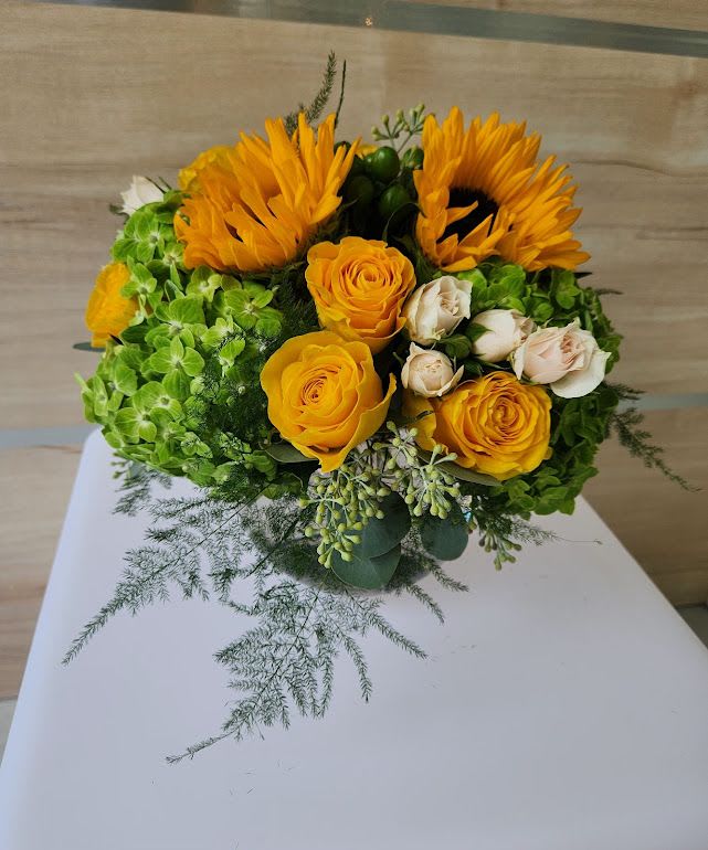 Round arrangement of yellow sunflowers and roses with green hydrangeas and cream spray roses.