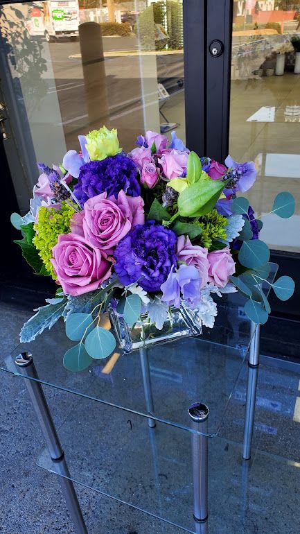 Low arrangement of pink roses and purple flowers in a square glass vase on a glass table.