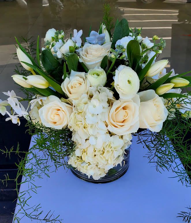 Low arrangement of ivory roses, tulips, orchids, and hydrangeas in a glass vase with trailing greenery