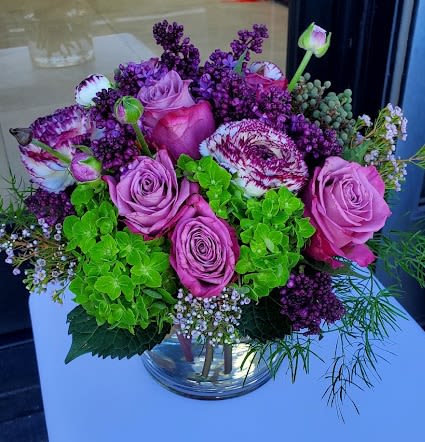 Compact arrangement of pink roses, purple blooms, and green hydrangeas in a clear glass vase