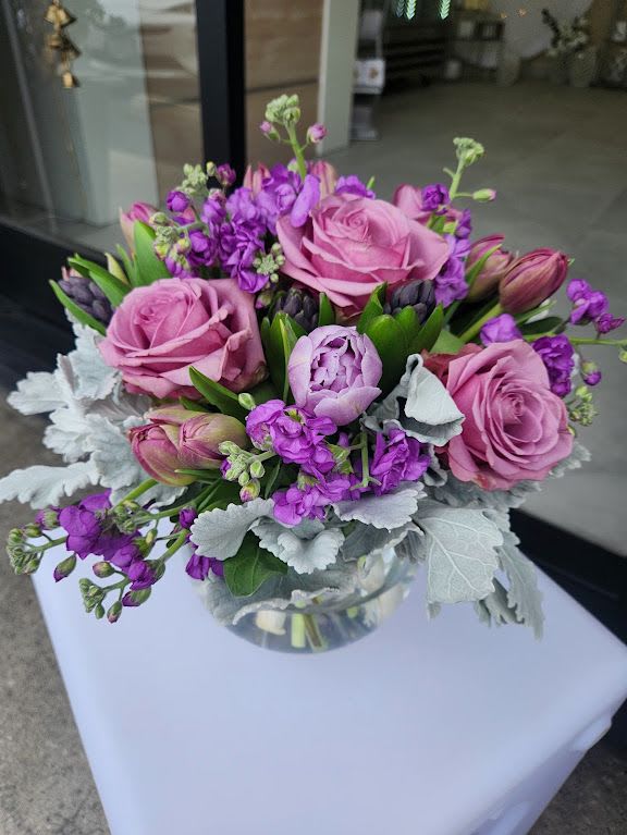 Round arrangement of pink roses and peony with purple blooms in a clear glass bowl vase