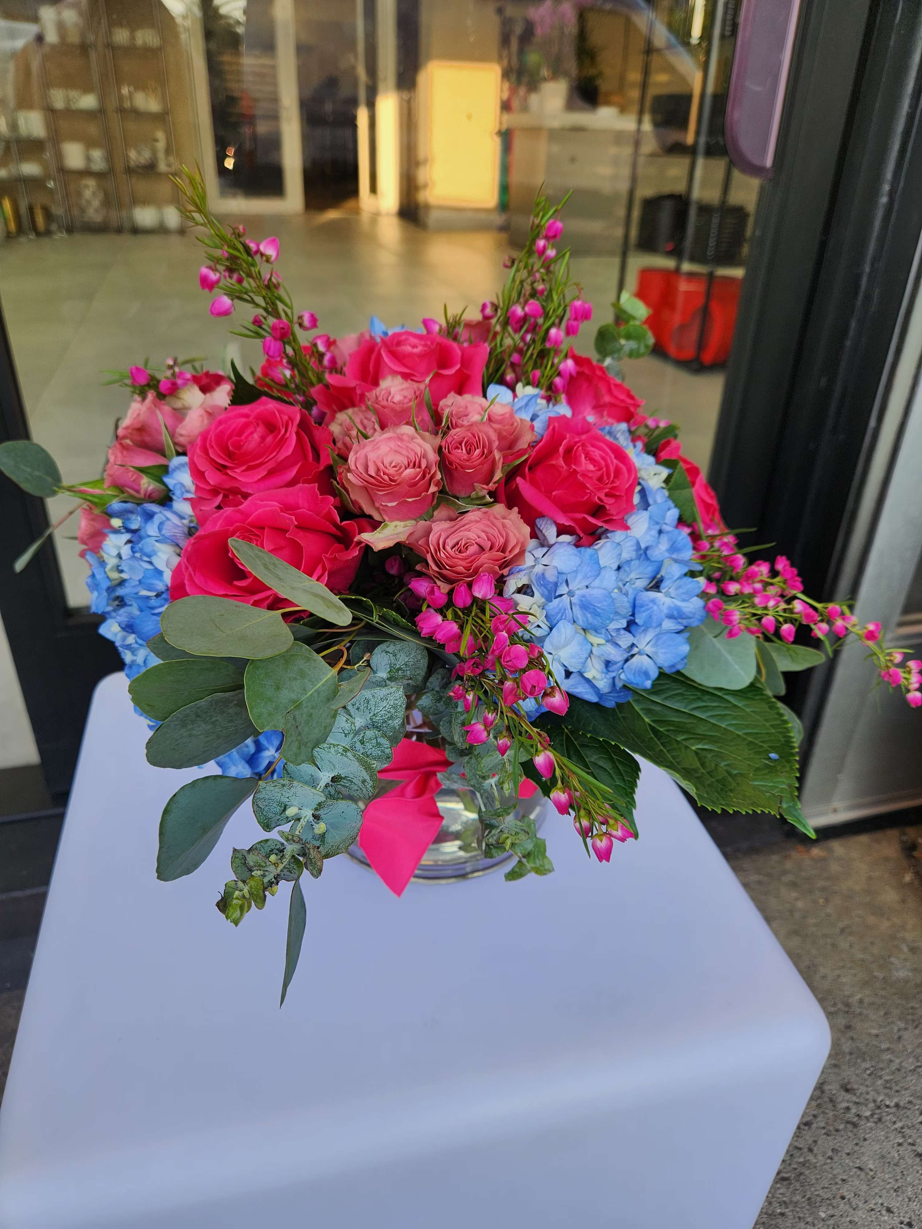 Low glass vase arrangement with hot pink roses and blue hydrangeas accented by small pink blossoms.