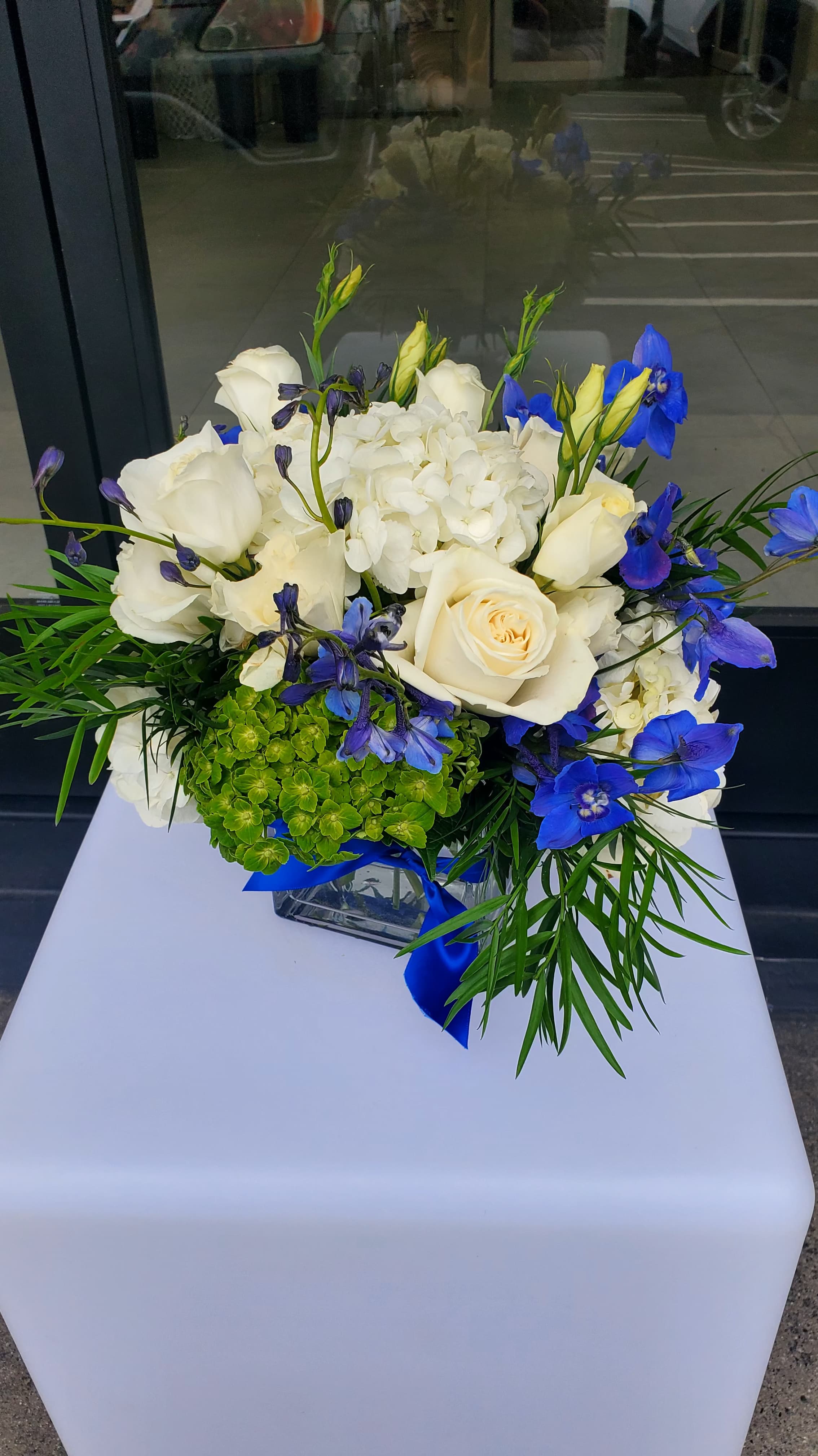 Low arrangement of white roses, hydrangeas, and bright blue flowers in a clear cube vase with a blue ribbon