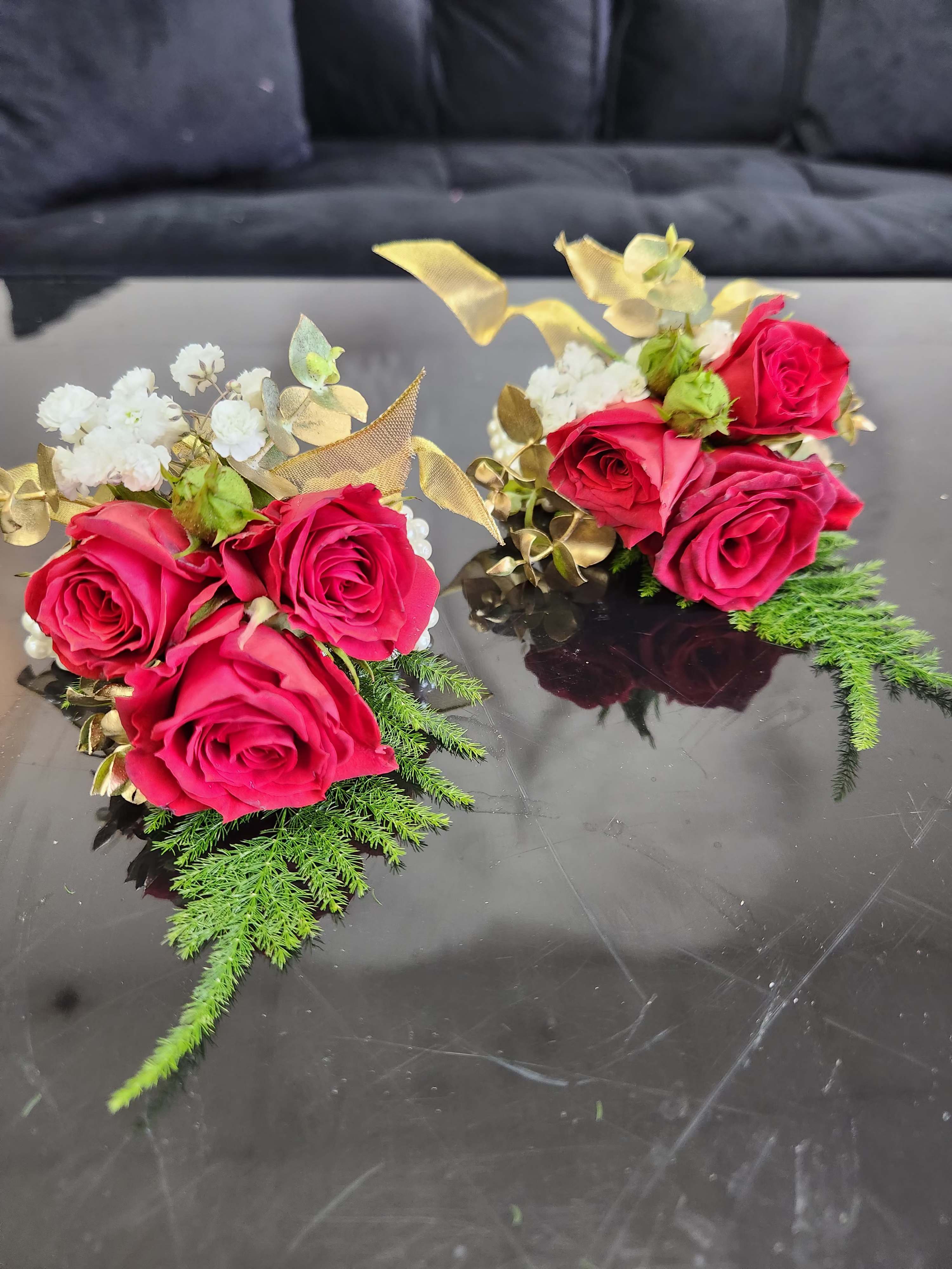 Pair of red rose and white accent flower wrist corsages with gold ribbon and fern accents on a reflective black surface.