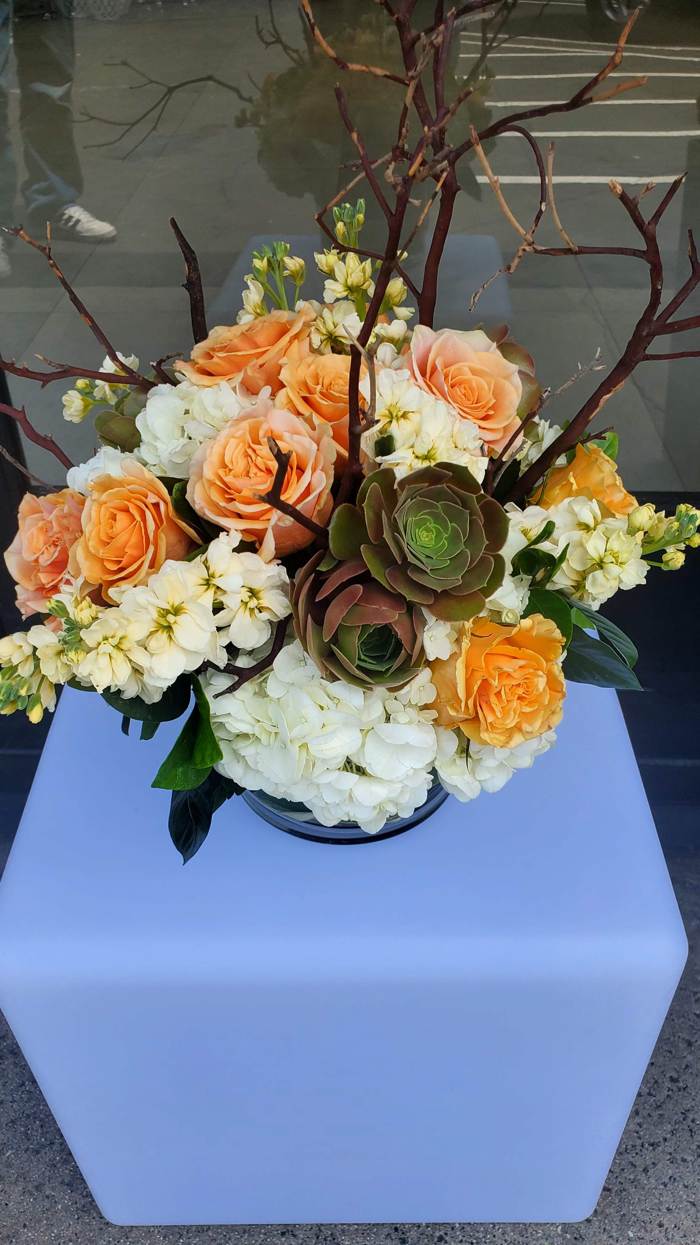 Peach roses, white hydrangeas, succulents, and branches arranged in a low glass vase.