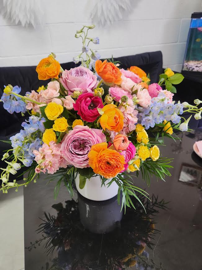 Bright mixed arrangement of pink, yellow, orange, and blue flowers in a white vase on a dark table