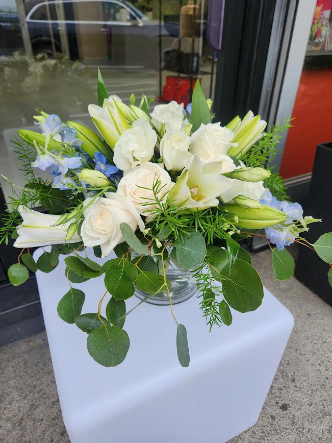 Low arrangement of white roses and lilies with light blue flowers in a clear glass bowl vase