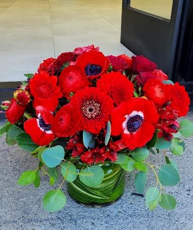 Low arrangement of vivid red flowers with dark centers in a leaf-wrapped glass vase