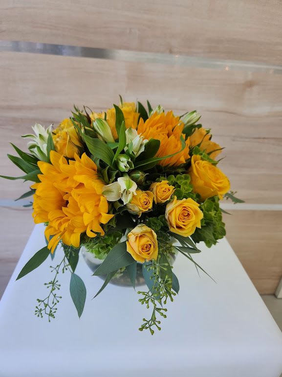 Low arrangement of yellow roses and yellow flowers in a round glass vase on a white table