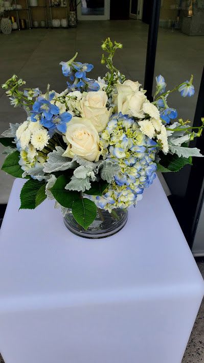Low round arrangement of cream roses, blue delphinium, hydrangeas, and white mums in a clear glass vase