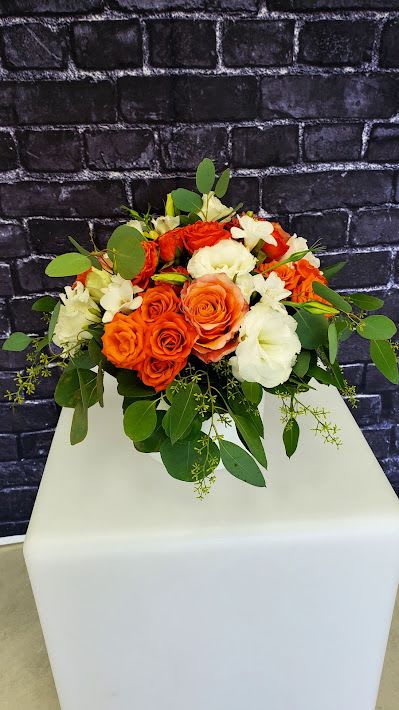 Low arrangement of orange roses and white blooms on a white pedestal with a dark brick backdrop