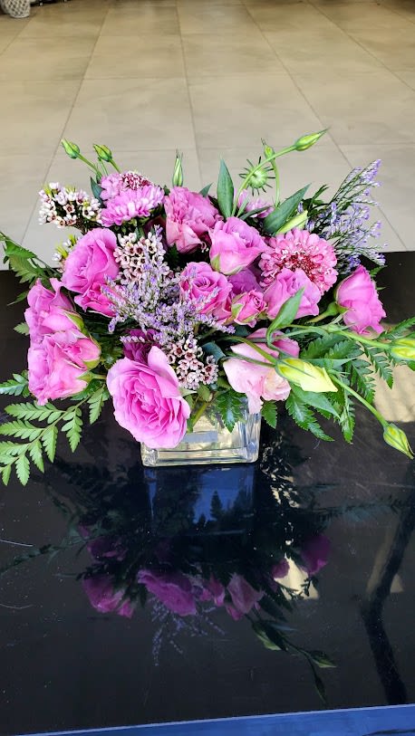 Low arrangement of pink roses and mixed purple flowers in a clear glass cube vase.
