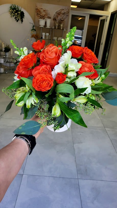 Arrangement of bright orange roses and white flowers with green foliage in a white round vase.
