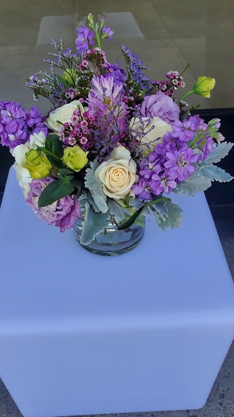 Low glass vase of cream roses and purple mixed flowers on a light blue cube.