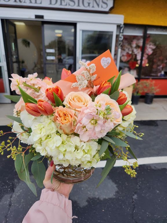 Low bouquet of peach roses, orange tulips, and white hydrangeas in a clear round vase with an orange card.