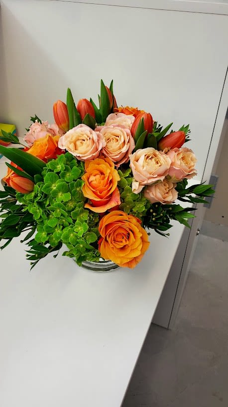Low arrangement of orange and peach roses with orange tulips and green hydrangea in a clear glass vase