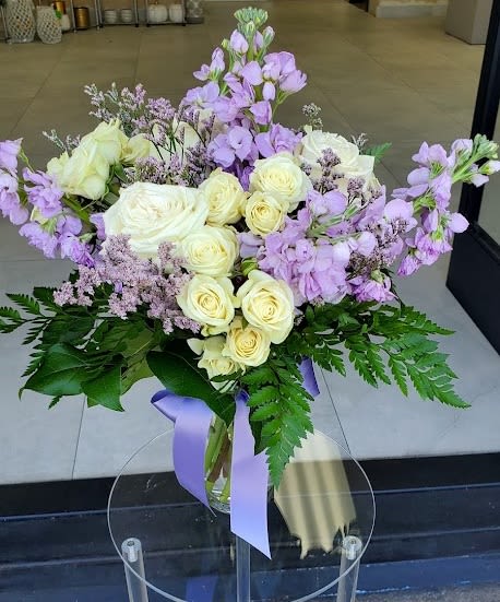 Vase arrangement of cream roses and light purple flowers with ferns and a lavender ribbon