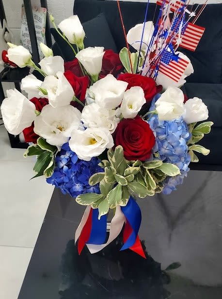 Patriotic red, white, and blue bouquet with roses and hydrangeas in a glass vase with American flags.