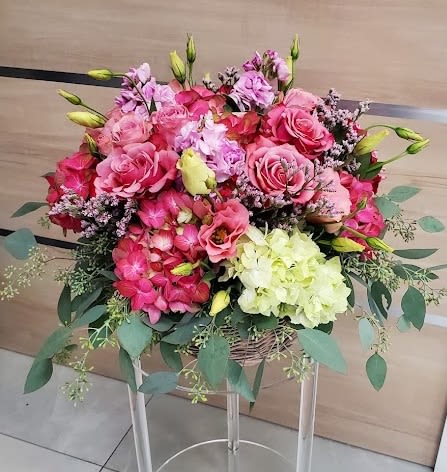Large pink and cream flower arrangement with roses and hydrangeas in a low basket on a clear stand