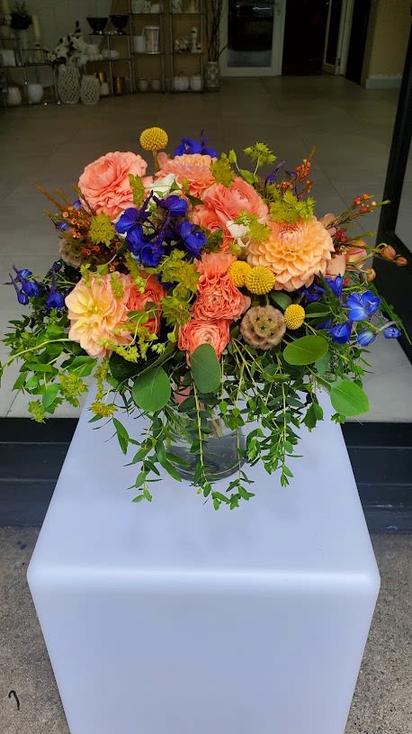 Large peach and blue mixed flower arrangement with trailing foliage in a clear glass vase.