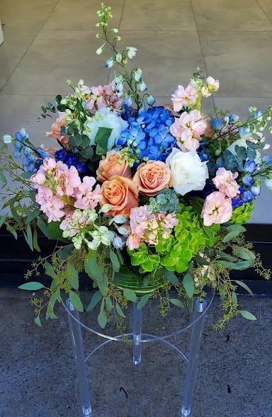 Large arrangement of blue hydrangeas, peach roses, and mixed blooms in a vase on a clear stand