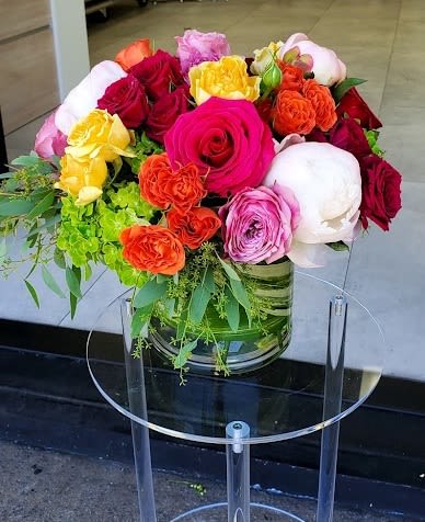 Low glass vase of bright roses, peonies, and spray roses in pink, red, orange, and yellow on a clear stand.