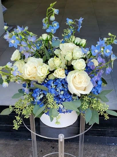 White roses and blue flowers arranged in a round white box on a clear stand