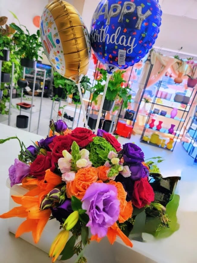 Colorful mixed flower arrangement with birthday balloons in a white rectangular container.