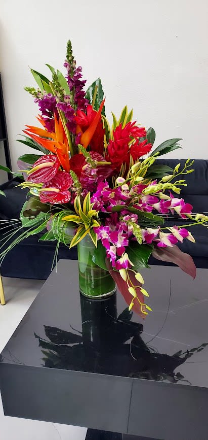 Tall tropical arrangement with purple orchids, red anthuriums and orange flowers in a clear glass vase