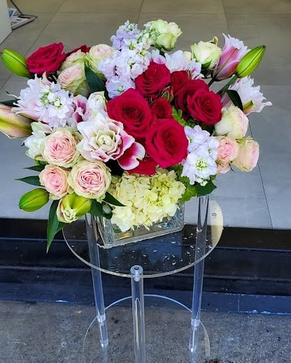 Low arrangement of red and pink roses, lilies, and light hydrangeas in a clear glass cube vase