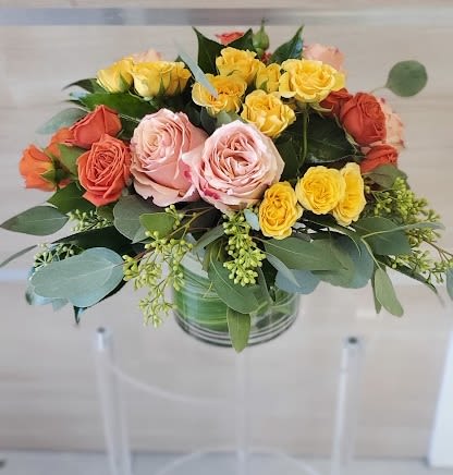 Round arrangement of yellow, peach, and orange roses in a clear glass cylinder vase