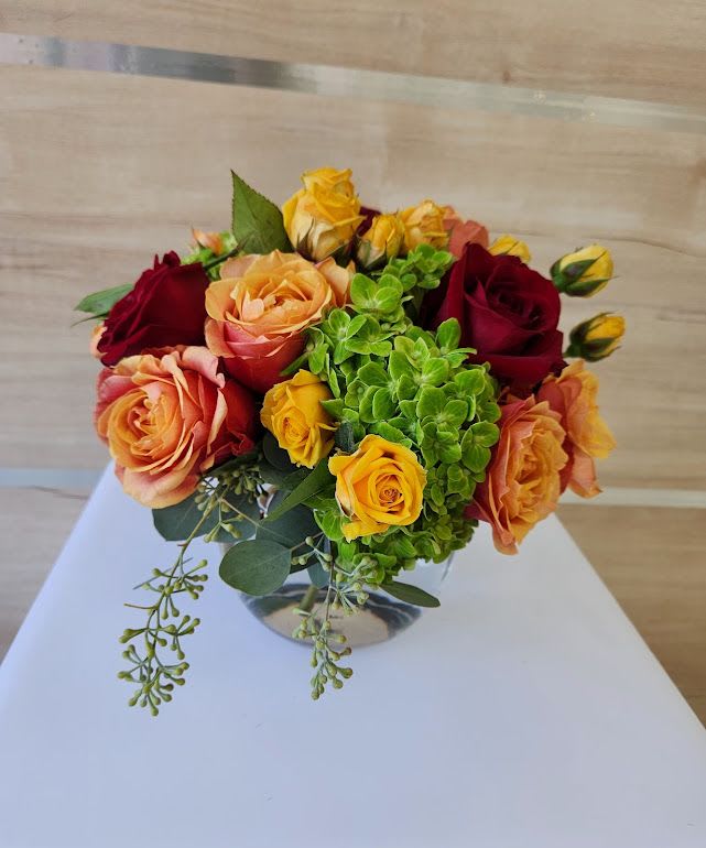 Low glass vase of orange, yellow, and red roses with bright green hydrangea