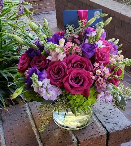 Low glass vase of fuchsia roses, purple blooms, and mixed pink flowers with a card tucked at the back.