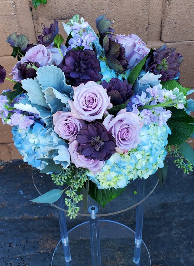Lavender roses and deep purple blooms arranged with blue hydrangeas in a rounded bouquet