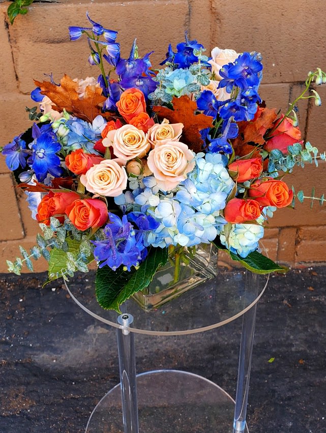 Arrangement of blue hydrangeas, blue delphinium, and orange and peach roses in a clear glass vase