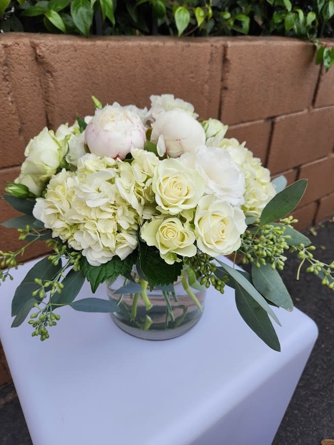 Low round arrangement of white roses, hydrangeas, and peonies in a clear glass vase