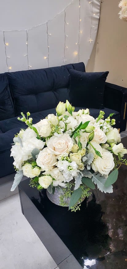 Low round arrangement of white and cream flowers in a silver container on a black table by a dark sofa