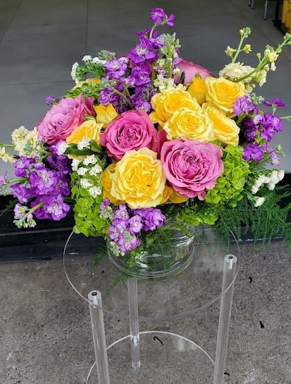 Arrangement of pink and yellow roses with purple stock and green hydrangeas in a clear glass vase.