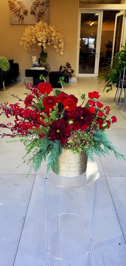 Low, lush arrangement of red roses and deep red flowers with evergreens in a gold container on a clear stand