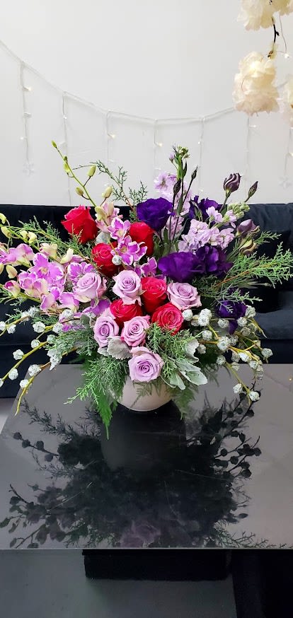 Low bowl arrangement of pink and red roses with purple blooms and orchids in a lush mixed design