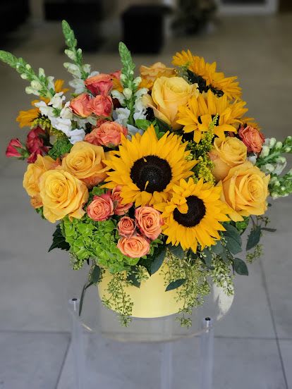 Arrangement of sunflowers, yellow and peach roses, and green hydrangea in a round yellow box.