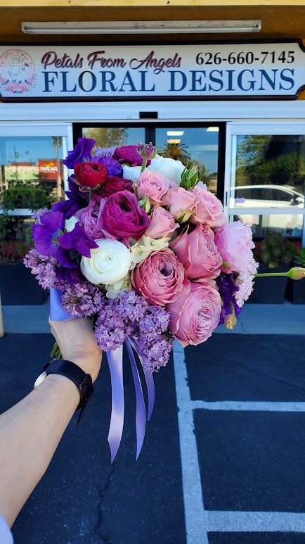 Hand-tied bouquet of pink roses, purple blooms, and white ranunculus with lavender ribbons.