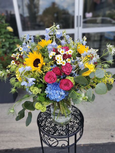 Summer Fun - Large mixed bouquet with sunflowers, hot pink roses, blue hydrangea, daisies in a tall clear glass vase.