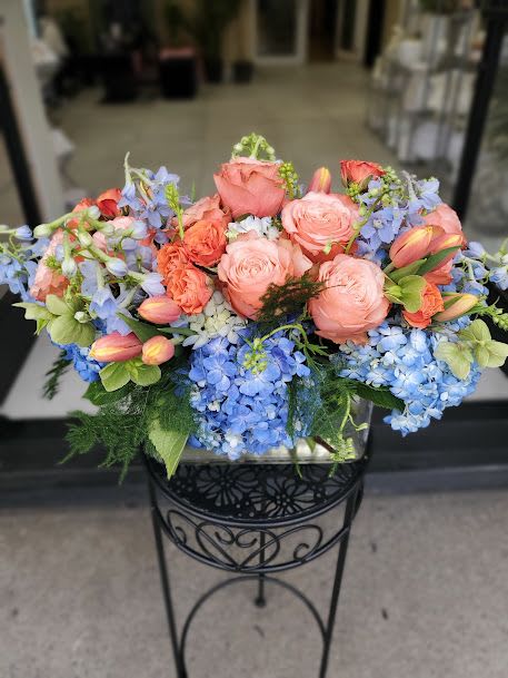 Peach and Blu - This lush, low-profile arrangement features a sophisticated blend of blush and coral roses, vibrant blue hydrangeas, and fresh tulips. The floral design is expertly displayed in a clear rectangular vase