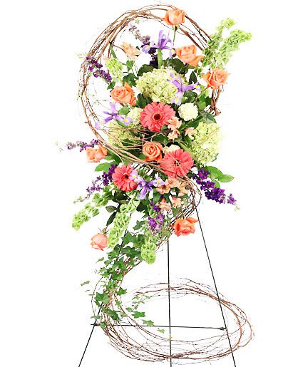 Bittersweet Dawn - Celebrate the brilliant life of someone near and dear to your heart with a Bittersweet Dawn standing spray that is perfect for any funeral, wake, or memorial service. Tropical tones, including soft greens and bright oranges, are excellent for highlighting the spirit of your loved one.  Includes Easel, Large Grapevine Wreath (Deconstructed), Large Wet Floral Foam Cage, Foliage: Ivy, Salal, Green Hydrangeas, Bells Of Ireland, Purple Larkspur, Orange Roses, Orange Alstroemeria, Coral Gerberas, Green Carnations, Purple Iris.