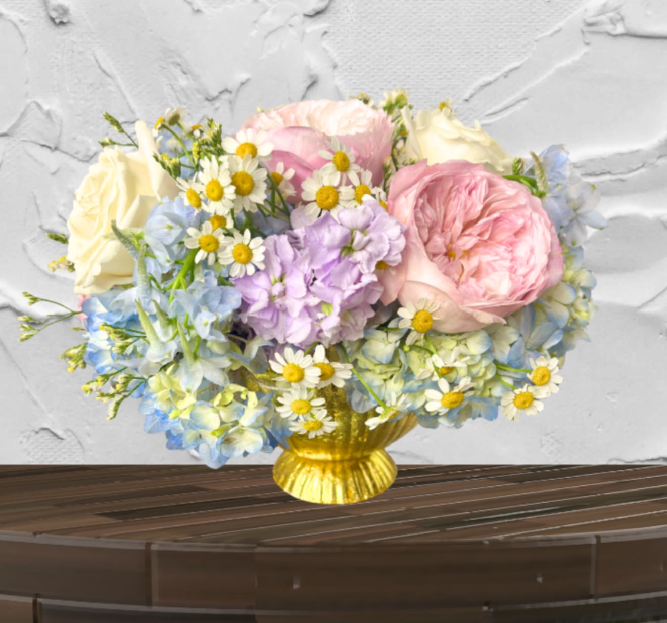 Golden Meadow Bouquet - The Golden Meadow Bouquet Sun-kissed charm meets garden elegance.  This whimsical, airy arrangement brings the brightness of a summer day into any room. Featuring lush pink Roses and ivory blooms, it is beautifully accented with sky-blue Delphinium, fragrant Lavender Stock, and cheerful Chamomile daisies.  Presented in a striking gold-toned pedestal vase, this arrangement offers a perfect balance of rustic charm and polished style.  Vibe: Joyful, breezy, and enchanting.  Key Blooms:  Roses, Chamomile, and Delphinium.  Perfect For: Birthdays, "thinking of you," or a bright kitchen centerpiece.