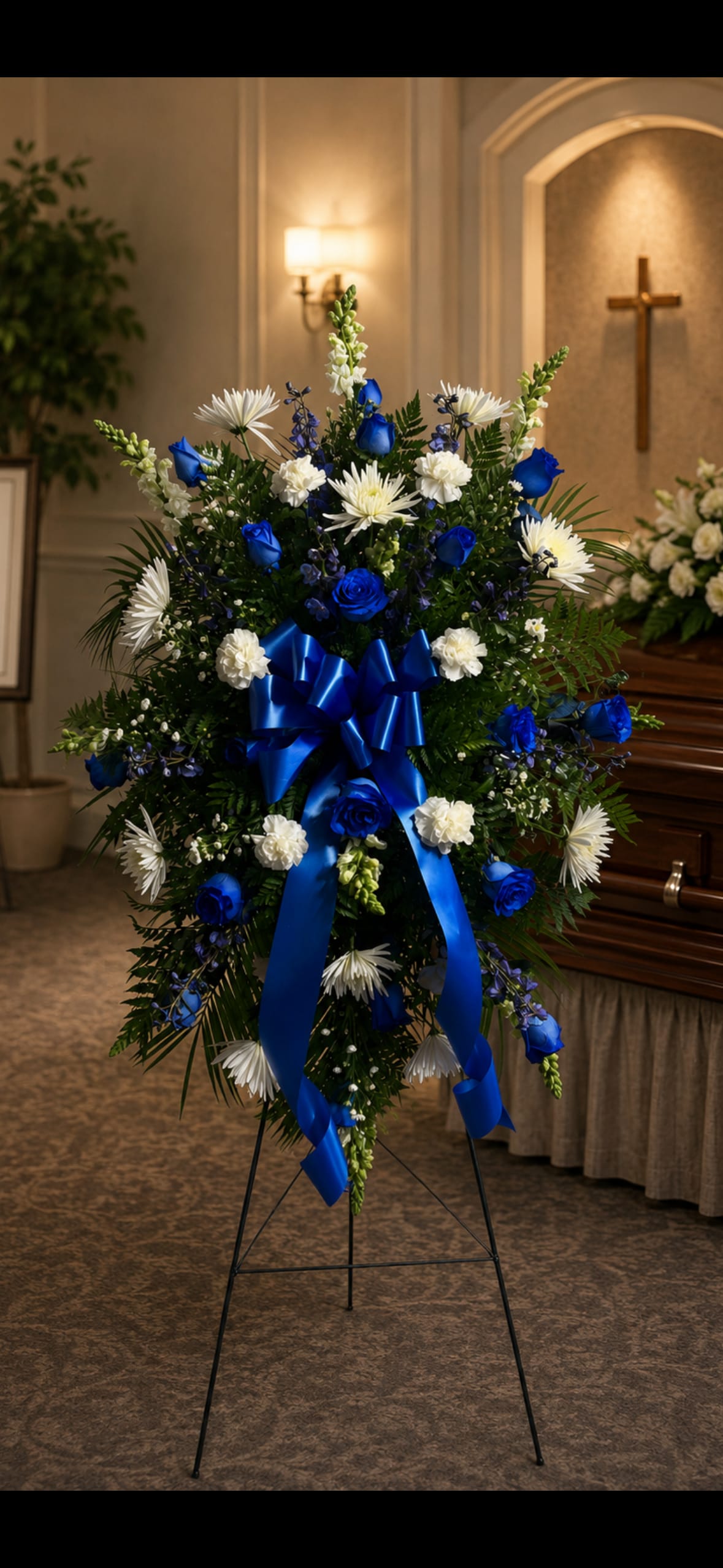 Blue And White Easel - Vivid royal-blue roses paired with crisp white carnations and white spiders with a white filler create a solemn, elegant standing spray perfect for sympathy or memorial services. This blue and white easel arrangement is designed to honor a life with formal balance and understated grace-the person you're remembering will be celebrated with dignity and thoughtful color.  A full, symmetrical shape of rounded carnations, spiky white snapdragons, and smaller blue accents sits against dark greenery on a sturdy metal easel stand. A wide satin blue ribbon bow with long streamers adds a ceremonial finish and the layered textures-rounded blooms, spiky whites, and delicate daisies-give the piece visual depth from every angle.  Choose this arrangement to convey respect and heartfelt sympathy at a funeral viewing or graveside service; it reads distinctly from across a room and photographs beautifully. Standard sizing fits most service spaces; if you need a larger or smaller version for a specific venue, contact us and we'll tailor the dimensions to suit the setting.