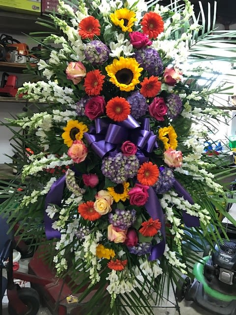 Large standing floral spray with sunflowers, gerbera daisies, roses, and purple ribbon