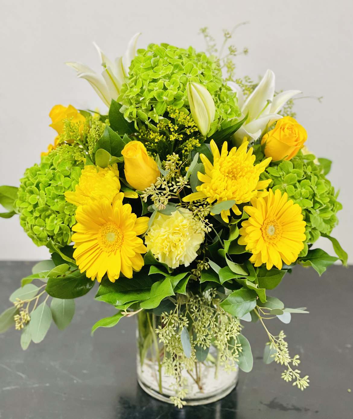 BF-E4 Springtime Splendor - Celebrate the vibrancy of nature with our stunning mix floral arrangement, meticulously crafted to enchant and delight. This exquisite ensemble features sunny yellow roses, cheerful gerbera daisies, lush green hydrangeas, and captivating Fuji mums, all elegantly arranged in a clear glass vase. 