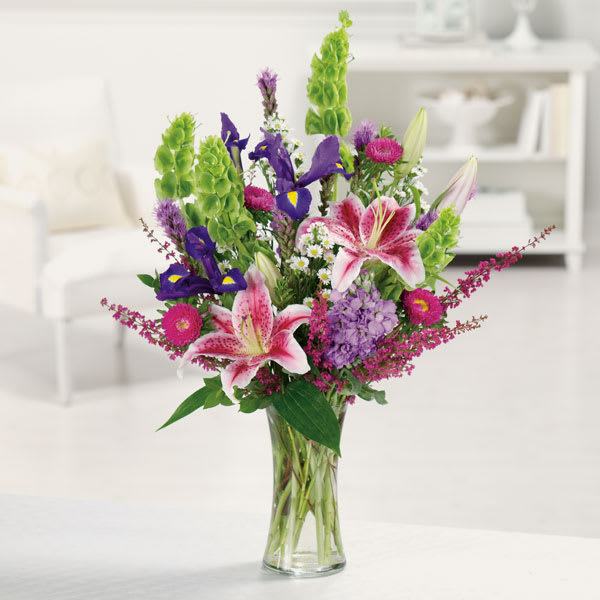 Mixed bouquet of pink lilies, purple irises, and green spikes in a glass vase